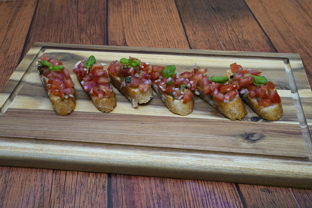 Mehrere Bruschetta liegen in einer Reihe auf einem Holzbrett und sind mit Tomaten und Basilikum belegt.