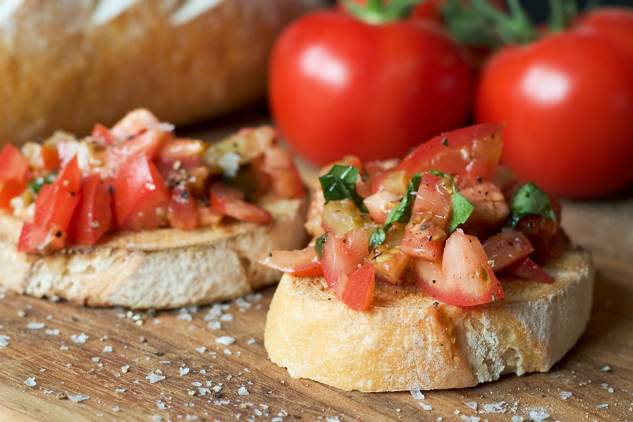 Zwei Bruschetta liegen auf einem Holzbrett, im Hintergrund stehen frische Tomaten und Brot.
