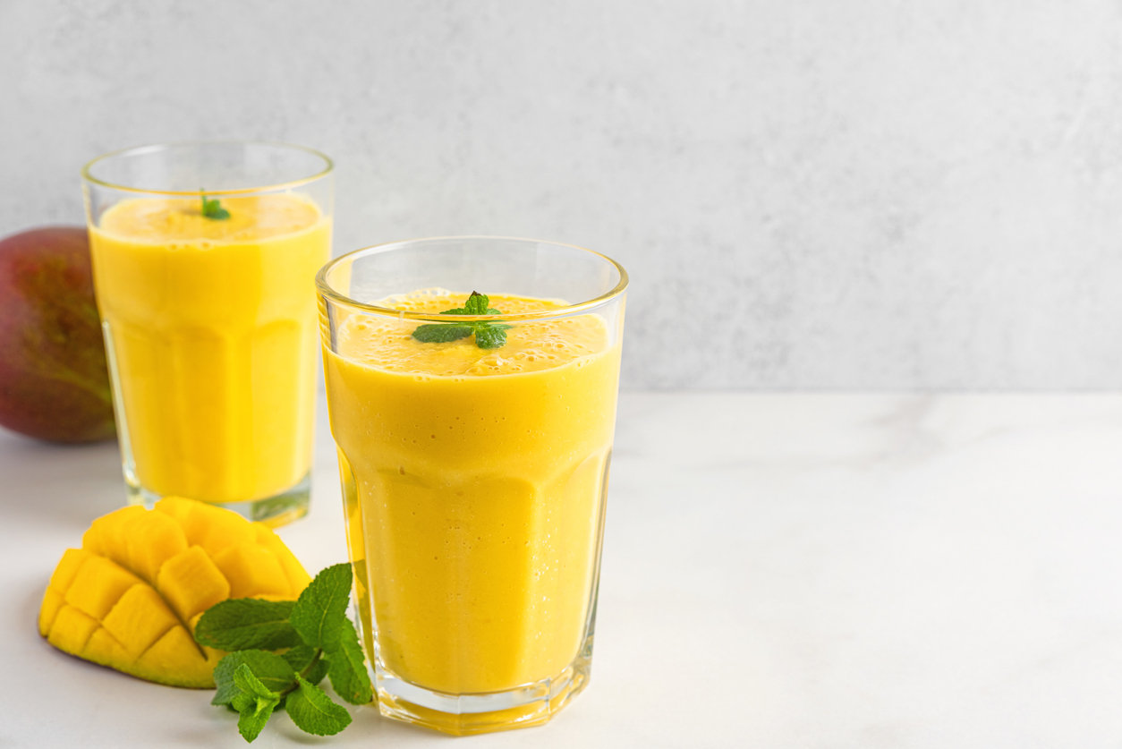 Zwei Gläser Mango Lassie stehen auf einem hellen Tisch, daneben liegen Mango und Minze.