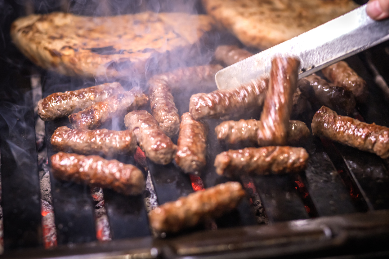 Eine Zange wendet Cevapcici auf dem Grill, während im Hintergrund Fladenbrot mitröstet.