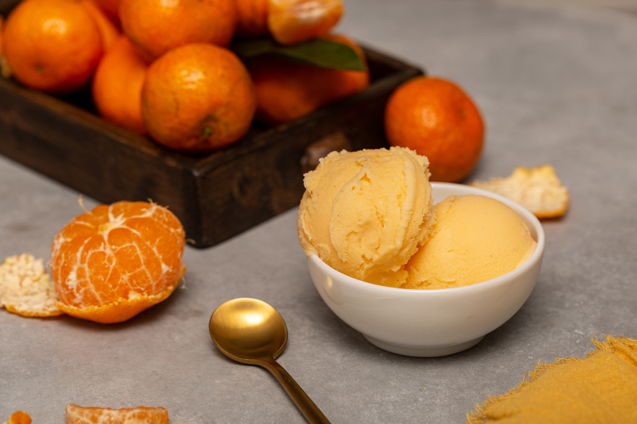 Zwei Kugeln Mandarineneis in einer Schale neben einer Holzkiste voller reifer Mandarinen und einem goldenen Löffel.
