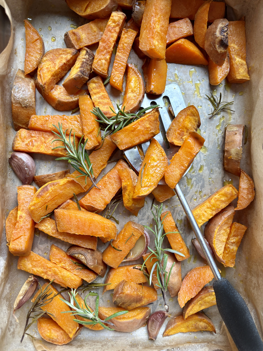 Eine Backform mit Süßkartoffeln steht neben einem Küchentuch und frischen Kräutern auf hellem Untergrund.