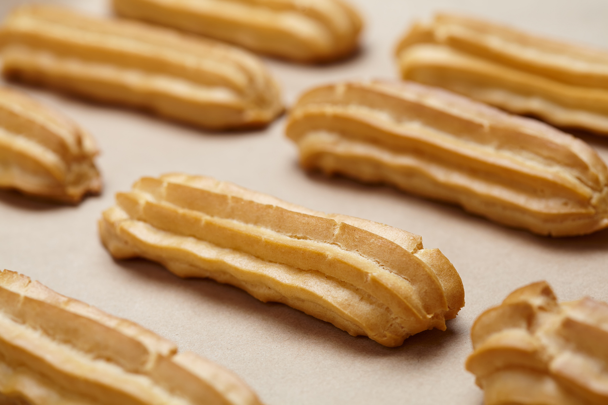 Eine perspektivische Nahaufnahme mehrerer Brandteig-Stangen auf Backpapier, wobei das vorderste Eclair scharf abgebildet ist.