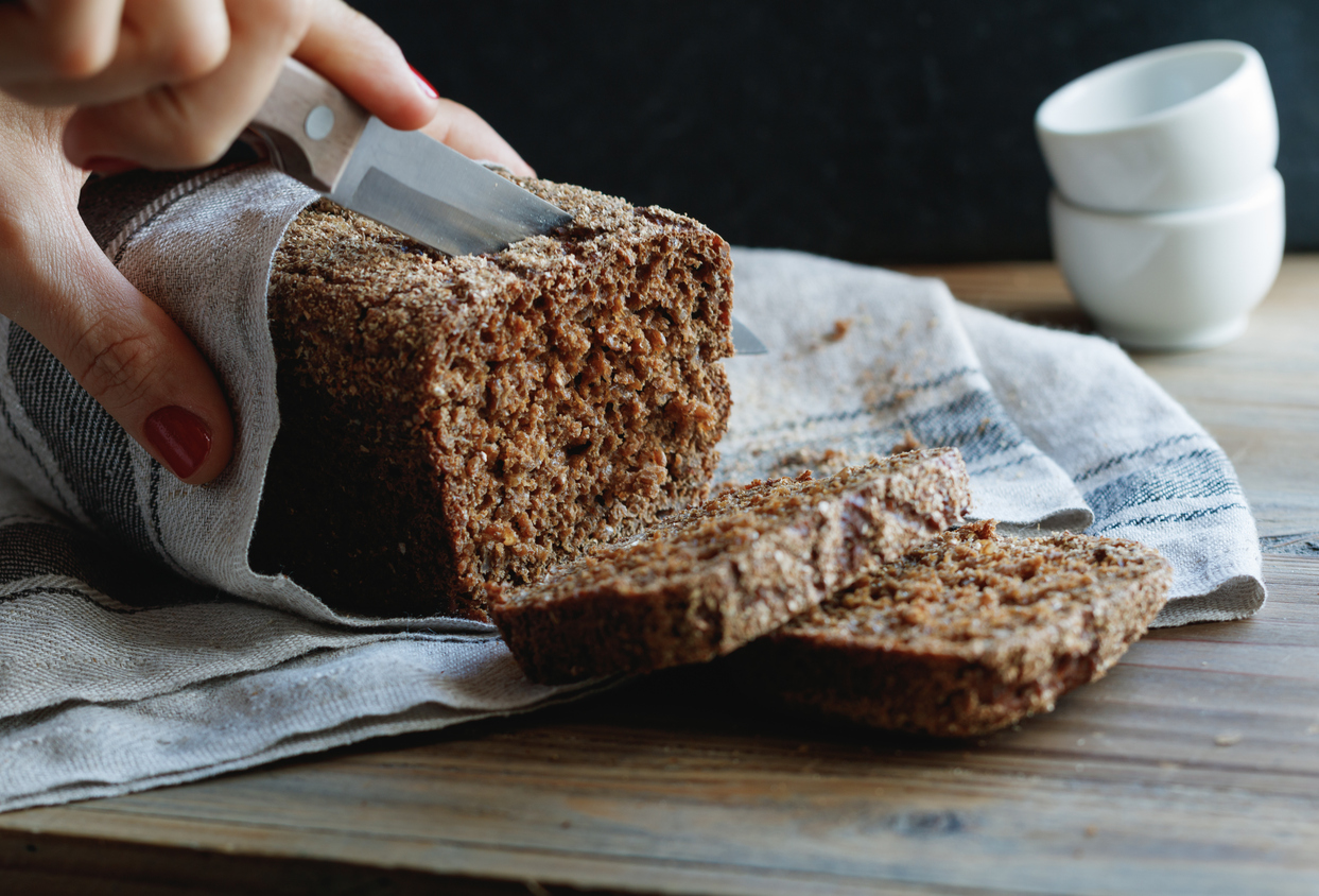 Eine Person schneidet mit einem Messer eine Scheibe von einem dunklen, grobporigen Roggenbrot ab.