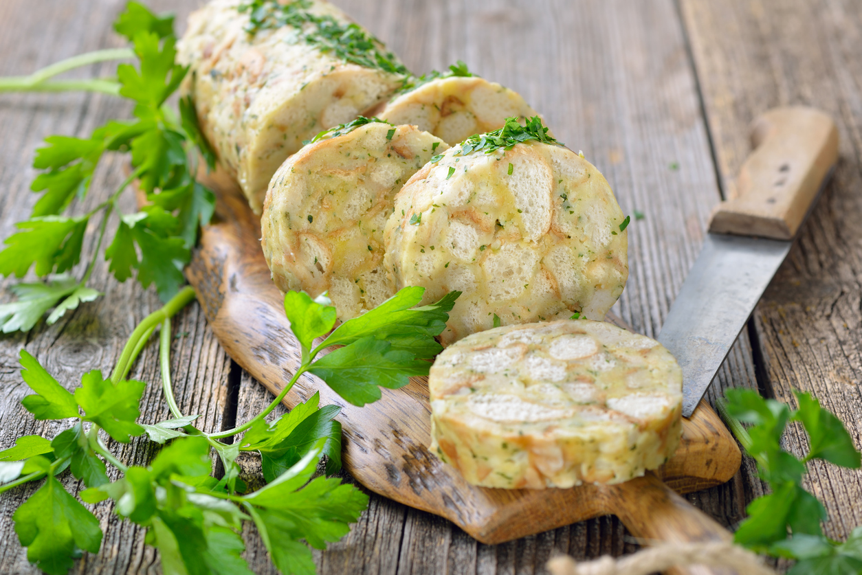 Geschnittene Semmelknödel liegen auf einem Holzbrett neben einem Messer und frischer Petersilie.
