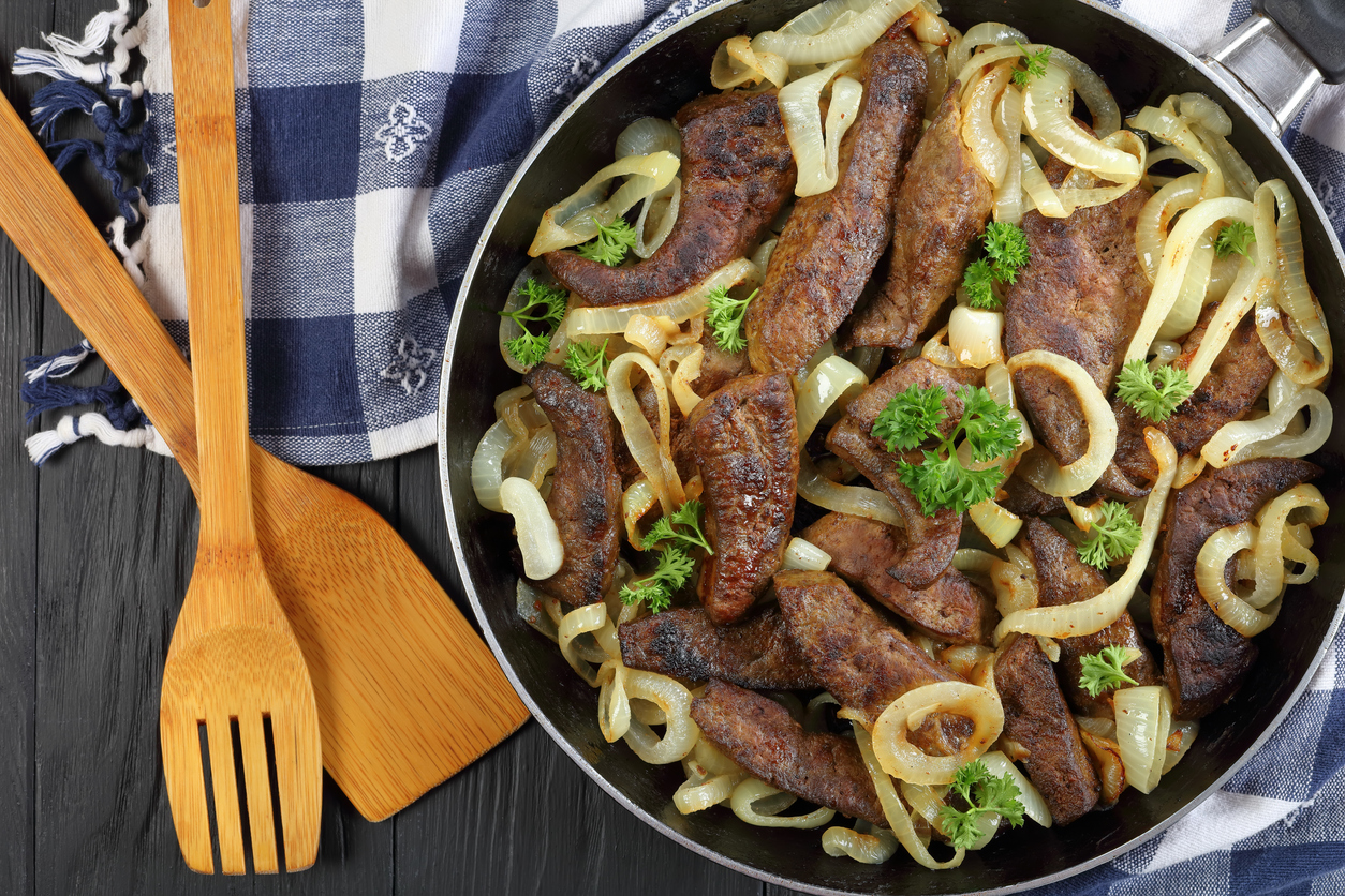 Gebratene Kalbsleber liegt mit Zwiebelringen und Petersilie in einer Pfanne auf dunklem Tisch.