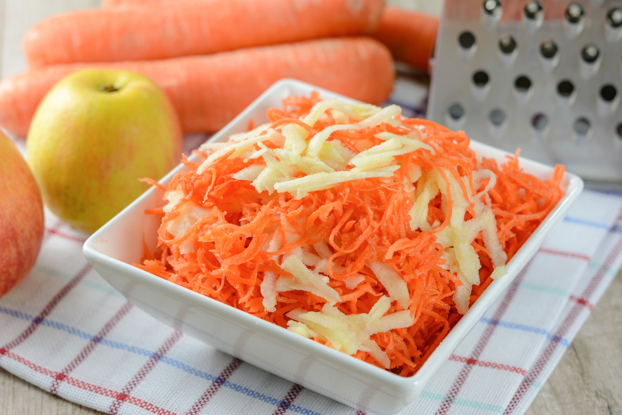 Karotten-Apfelsalat liegt in einer weißen Schale, daneben sind Äpfel und Karotten zu sehen.