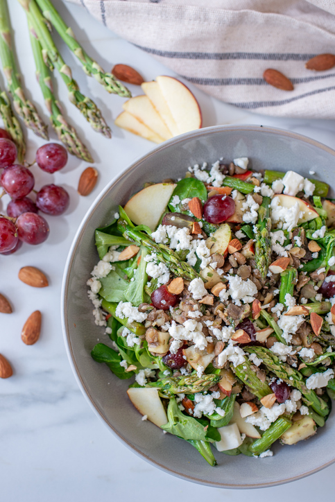Eine Schale mit grünem Spargelsalat, Trauben, Apfelstücken, Mandeln und weißem Käse steht auf einer hellen Fläche.