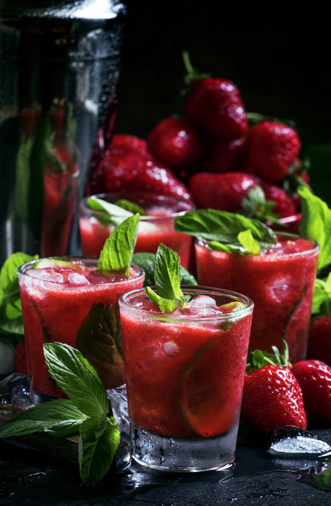 Mehrere Gläser Erdbeercaipirinha mit Minzblättern und Eiswürfeln stehen vor einem Hintergrund aus Erdbeeren und Barzubehör.