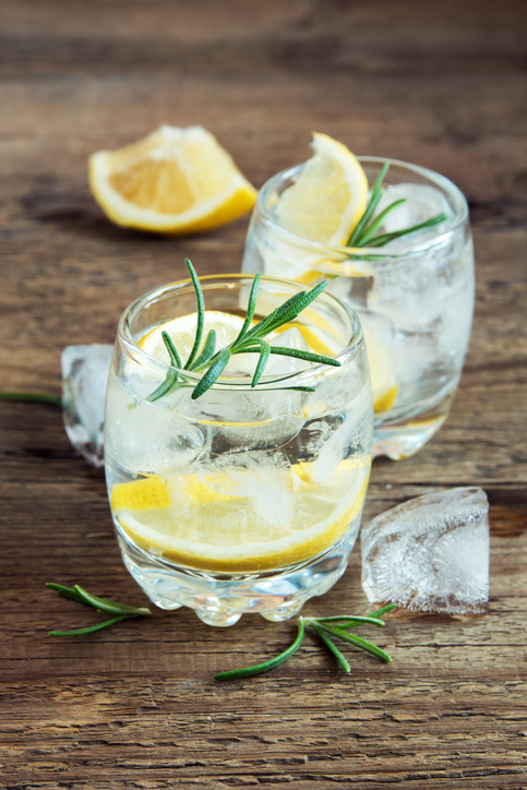 Nahaufnahme von zwei Gläsern Gin Tonic mit Eiswürfeln, frischen Zitronenscheiben und einem Rosmarinzweig auf einem rustikalen Holztisch.