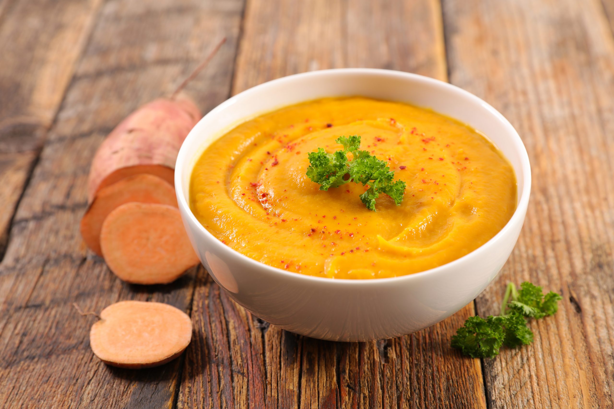 Eine Schale mit cremiger, orangefarbener Süßkartoffelsuppe, garniert mit Kürbiskernen, Petersilie und einem Klecks Sahne.