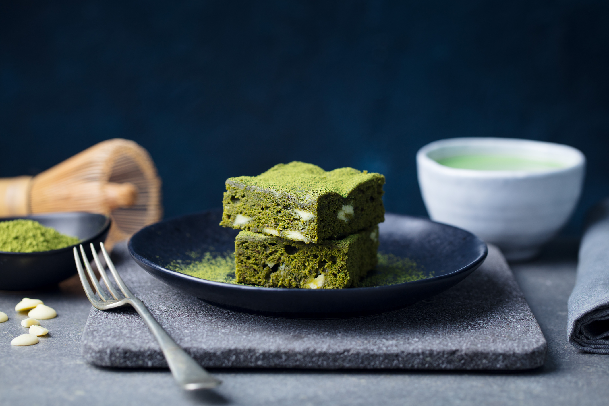 Zwei übereinander gestapelte, leuchtend grüne Matcha-Brownie-Stücke liegen auf einem dunklen Teller, bestäubt mit feinem Matcha-Pulver, begleitet von einem Matchatee und Backutensilien im Hintergrund.