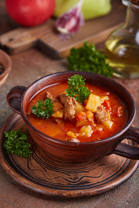 Gulaschsuppe in einer braunen Tonschale mit Fleischstücken, Kartoffeln, Gemüse und frischer Petersilie.